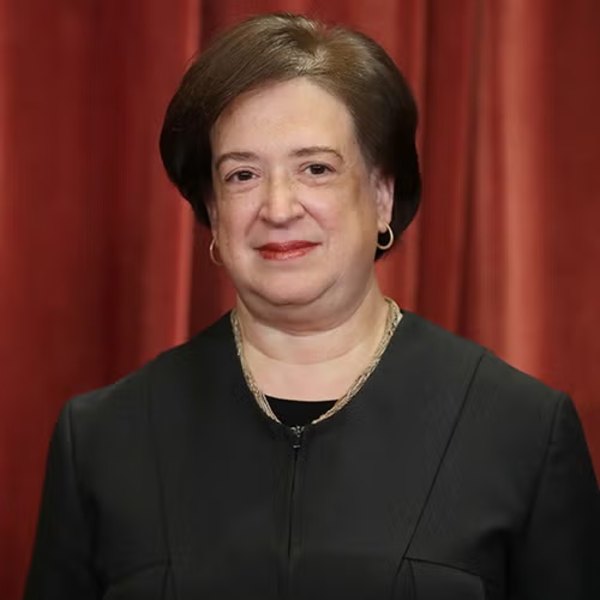 Justice Elena Kagan wearing a black judicial robe, posed before red curtains.
